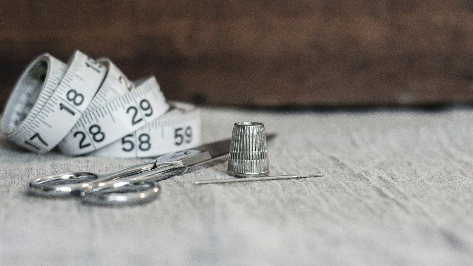 Sammenrullet målebånd, en liten saks, et fingerbøl og en nål på grå linduk