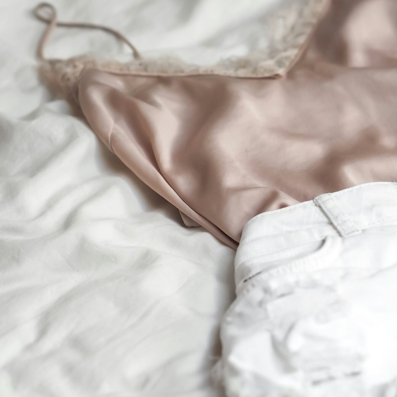 Pudderrosa satengtopp med blondekant på et hvitt laken, ved siden av et par hvite jeans
