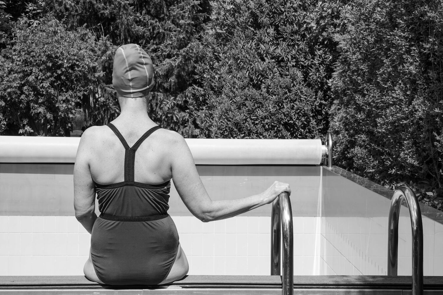 Svart-hvitt bilde av en kvinne i badehette og badedrakt som sitter på bassengkanten, sett bakfra