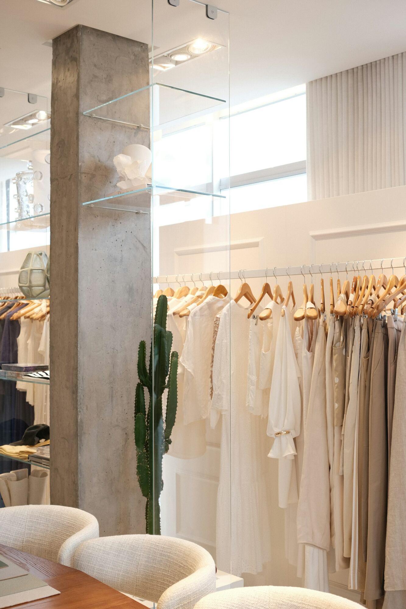 Lyst butikklokale med kjoler og bluser i hvitt og beige på stativ, glasshyller og en kaktus