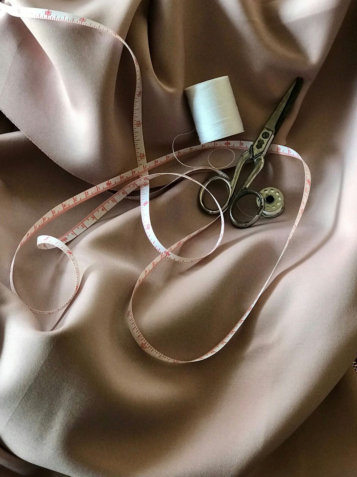 Målebånd som ligger over draperte stoffer i rosa og beige, med en trådsnelle og en saks